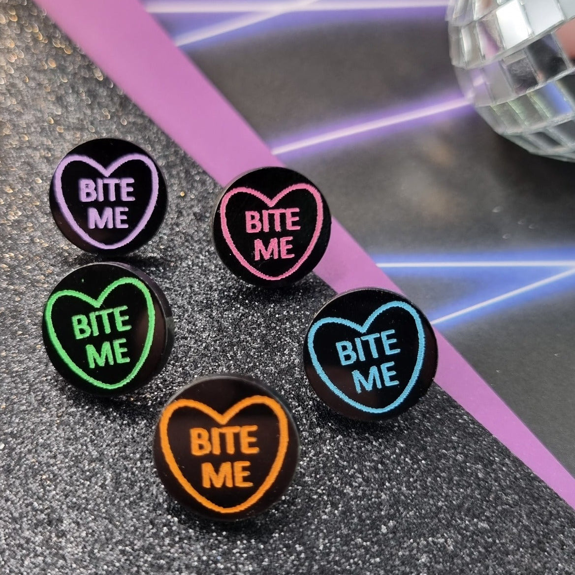 Round black stud earrings with neon heart outline and Bite Me lettering